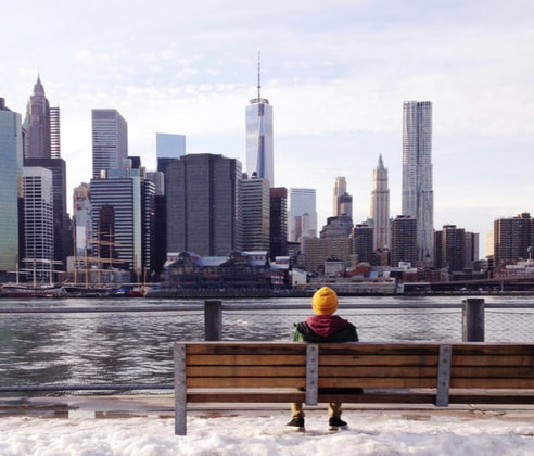 a person in a bright and warm hat sits on a park bench looking over the river to the NYC skyline. There is snow on the ground.
